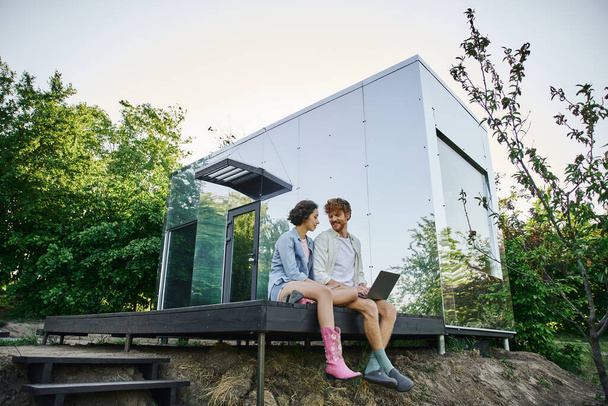 jovem casal multiétnico sentado no alpendre com laptop perto de casa de vidro moderno no campo - Foto, Imagem