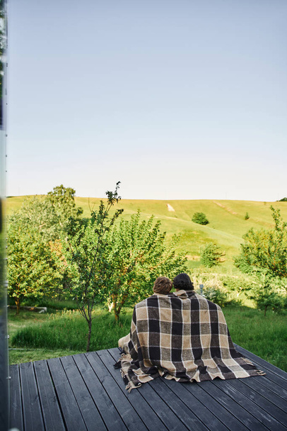 back view of young couple sitting on wooden porch under plaid blanket and enjoying scenic landscape - Фото, изображение
