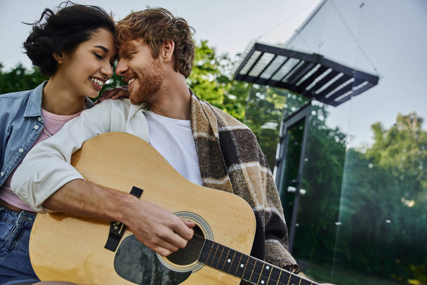 happy redhead man sitting face to face with asian girlfriend and playing guitar near glass house - Fotografie, Obrázek