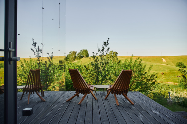 sillas de madera y mesa de centro en el porche cerca de la moderna casa de cristal con vista al paisaje escénico - Foto, Imagen