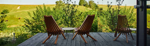 sillas de madera y mesa de centro en el porche con vista al paisaje escénico cerca de la construcción de vidrio, pancarta - Foto, Imagen
