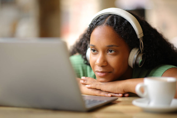 Black woman wearing headphone watching media on laptop in a restaurant terrace - Fotó, kép