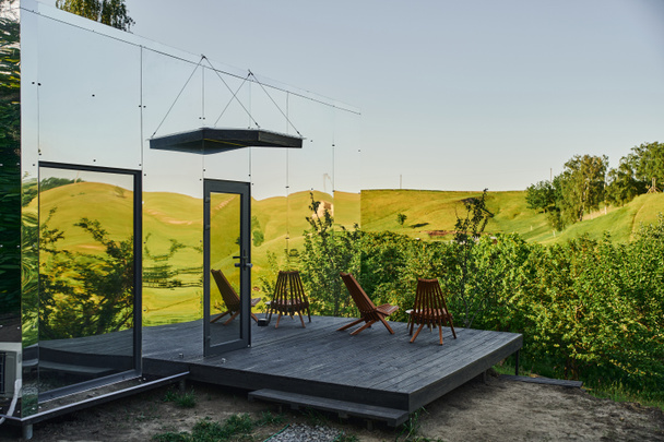 casa de cristal ecológica con sillas en el porche de madera y vista al pintoresco paisaje rural - Foto, Imagen