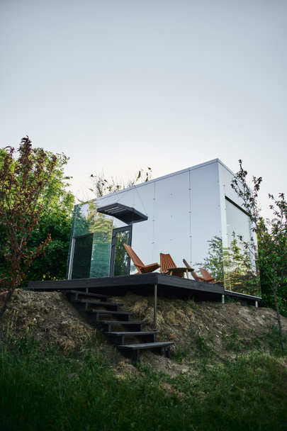 moderna casa de cristal ecológico con escaleras de madera y silla en el porche, entorno rural tranquilo - Foto, Imagen
