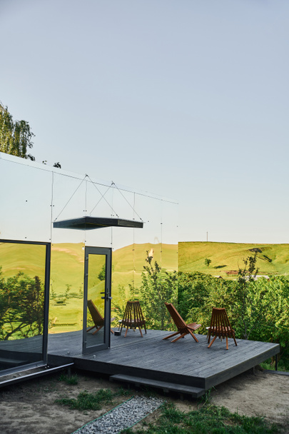 umweltfreundliches Glashaus mit Holzveranda in der Nähe grüner Hügel unter blauem Himmel in malerischer Landschaft - Foto, Bild