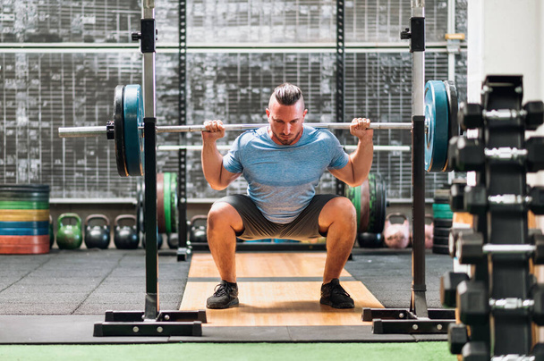 Focused male athlete in sportswear looking down while standing on gym floor lifting heavy barbell and performing back squat series - Photo, Image