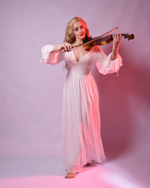 Full length portrait of beautiful blonde model wearing elegant white halloween gown, historical fantasy character.  facing backwards holding a violin musical instrument, isolated on studio background. - Photo, Image