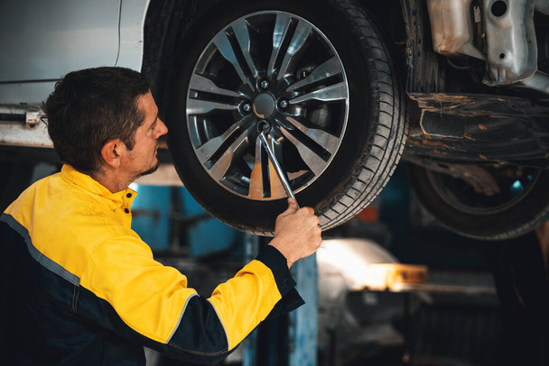 Hardworking mechanic changing car wheel under car lifting station. Automotive service worker changing leaking rubber tire in concept of professional car care and maintenance. Oxus - Photo, Image