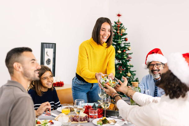 Wielopokoleniowa duża rodzina ciesząca się świąteczną kolacją w domu. Szczęśliwa młoda kobieta serwująca sałatkę przy stole podczas świąt Bożego Narodzenia - Zdjęcie, obraz