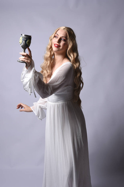 Close up portrait of beautiful blonde model wearing elegant  white halloween gown, a historical fantasy character.  Holding wine goblet, isolated on audio background. - Photo, Image
