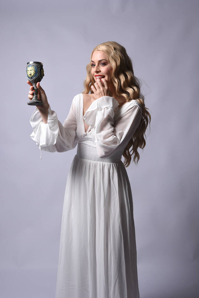 Close up portrait of beautiful blonde model wearing elegant  white halloween gown, a historical fantasy character.  Holding wine goblet, isolated on audio background. - Zdjęcie, obraz
