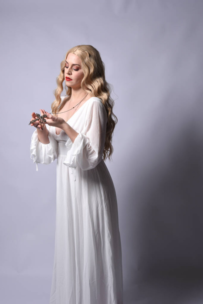 Close up portrait of beautiful blonde model wearing elegant  white halloween gown, a historical fantasy character.  Holding crucifix cross necklace, isolated on studio background. - Foto, Imagem