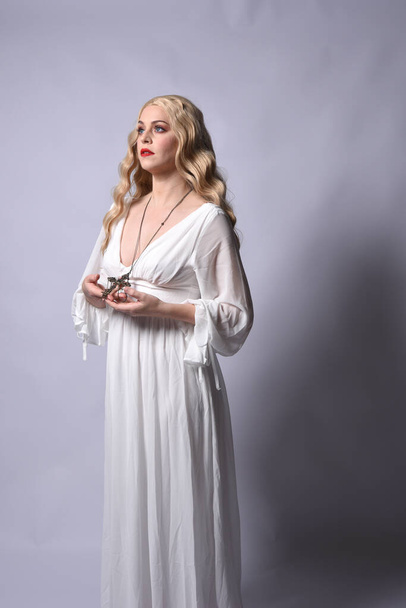 Close up portrait of beautiful blonde model wearing elegant  white halloween gown, a historical fantasy character.  Holding crucifix cross necklace, isolated on studio background. - Photo, Image