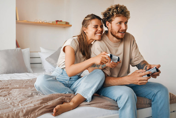 Jeune couple amoureux jouant à des jeux vidéo tout en étant assis sur le canapé dans la chambre à coucher, s'amuser et profiter l'un de l'autre le week-end. - Photo, image