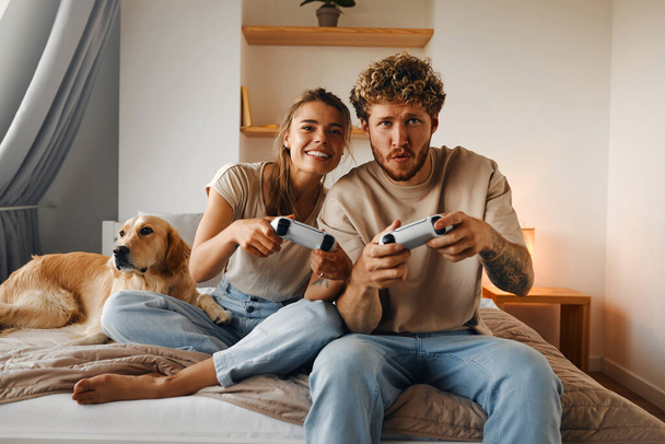 Jong stel verliefd spelen videospelletjes terwijl zitten op de bank in de slaapkamer, plezier hebben en genieten van elkaar in het weekend. - Foto, afbeelding