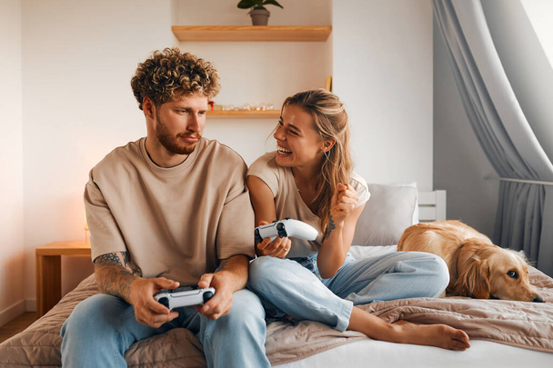 Jeune couple amoureux jouant à des jeux vidéo tout en étant assis sur le canapé dans la chambre à coucher, s'amuser et profiter l'un de l'autre le week-end. - Photo, image