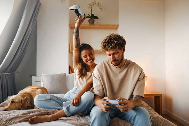 Giovane coppia innamorata di giocare ai videogiochi mentre si siede sul divano in camera da letto, divertirsi e godersi l'un l'altro nei loro fine settimana. - Foto, immagini
