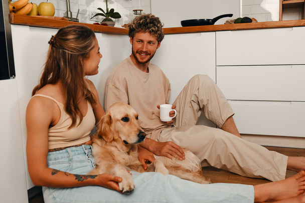 Ένα νεαρό παντρεμένο ζευγάρι ερωτευμένο να κάθεται στο πάτωμα της κουζίνας πίνοντας καφέ το πρωί με τον αγαπημένο τους σκύλο, περνώντας ρομαντικά χρόνο μαζί. - Φωτογραφία, εικόνα
