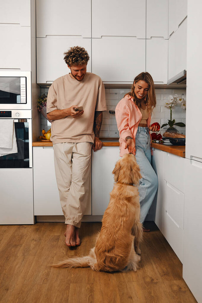 Νεαρό ερωτευμένο παντρεμένο ζευγάρι παίρνει πρωινό το πρωί μαζί στην κουζίνα με το αγαπημένο τους σκυλί σε ένα άνετο σπίτι. - Φωτογραφία, εικόνα