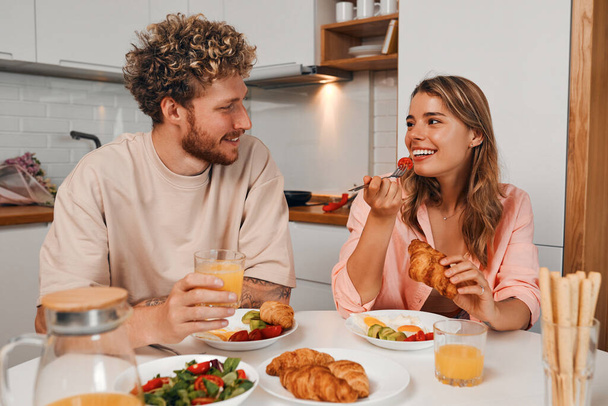 Een jong getrouwd stel dat 's ochtends in de keuken ontbeten in een gezellig huis op hun vrije dag terwijl ze romantisch samen tijd doorbrengen. - Foto, afbeelding