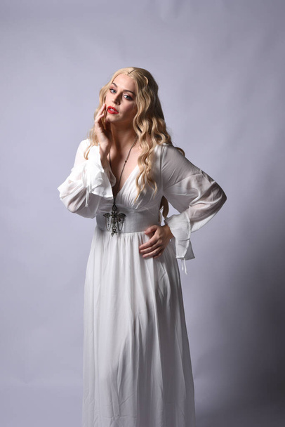 Close up portrait of beautiful blonde model wearing elegant  white halloween gown, a historical fantasy character.  Holding crucifix cross necklace, isolated on studio background. - Φωτογραφία, εικόνα