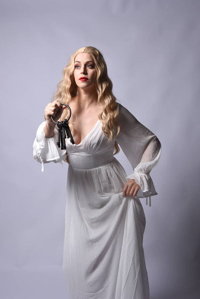 Close up portrait of beautiful blonde model wearing elegant  white halloween gown, a historical fantasy character. Holding old key chain, isolated on studio background. - Photo, Image