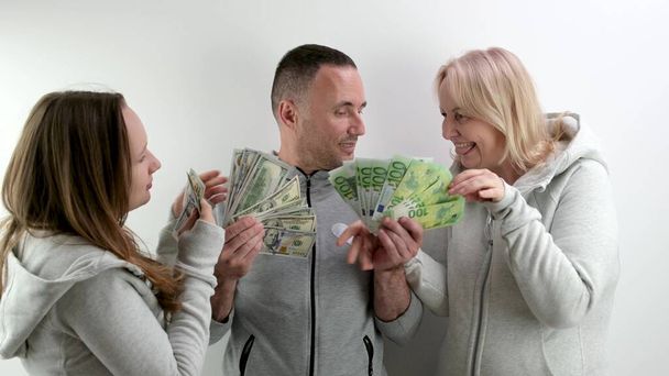 man father and husband stand with money rejoicing in life daughter and wife come and take all money man spreads his arms to the sides frustrated remains alone tracksuits. Cheerful family dollars euros - Photo, Image