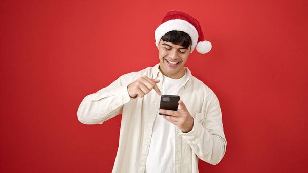 Молодий іспаномовний чоловік у різдвяній капелюсі вказує на смартфон над ізольованим червоним тлом - Фото, зображення