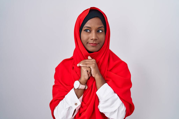 Młoda arabka w tradycyjnym islamskim szaliku śmieje się nerwowo i podniecona dłońmi na podbródku patrząc z boku  - Zdjęcie, obraz