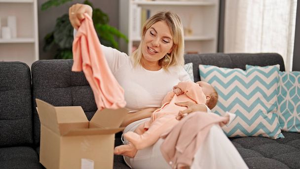 Mother and daughter holding baby clothes of package sitting together at home - Foto, afbeelding