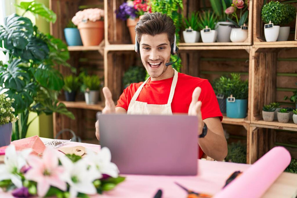 Young hispanic man working at florist shop doing video call looking at the camera smiling with open arms for hug. cheerful expression embracing happiness.  - Фото, изображение