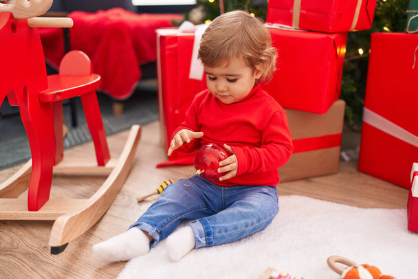 Imádnivaló spanyol kisbaba kezében karácsonyi labda dekoráció ül a padlón otthon - Fotó, kép