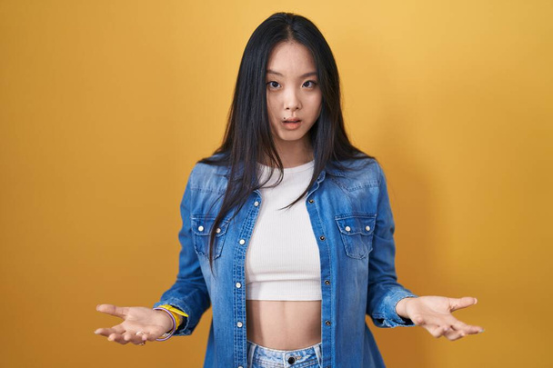 Fiatal ázsiai nő áll a sárga háttér tanácstalan és zavaros a nyitott karok, fogalmam sincs koncepció.  - Fotó, kép