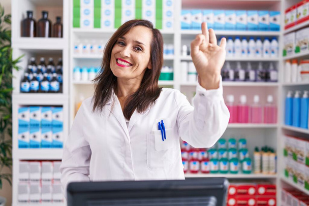 Μεσήλικη μελαχρινή γυναίκα που εργάζεται στο φαρμακείο χαμογελώντας θετικά κάνει ok σημάδι με το χέρι και τα δάχτυλα. επιτυχής έκφραση.  - Φωτογραφία, εικόνα
