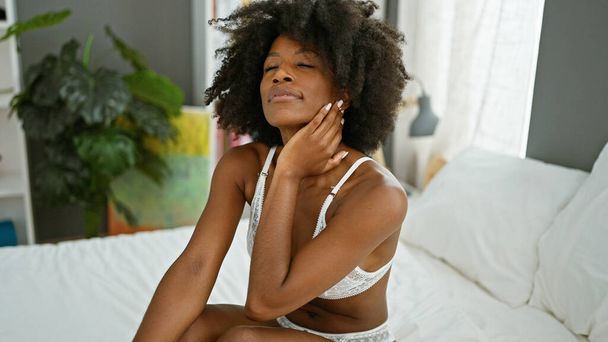 Африканская американка, сидящая на кровати, выглядит сексуально с серьезным лицом в спальне - Фото, изображение