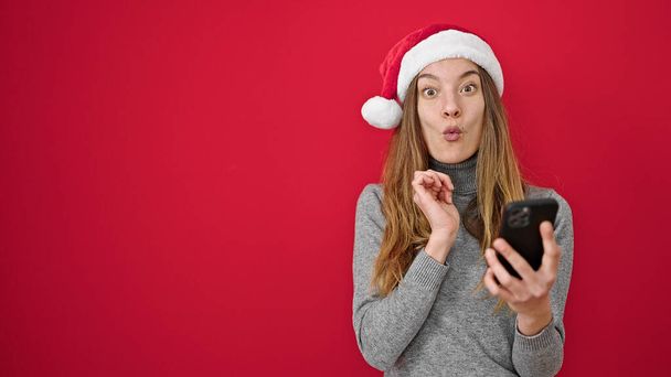 Mujer caucásica joven que usa sombrero de Navidad con teléfono inteligente con expresión sorpresa sobre fondo rojo aislado - Foto, imagen