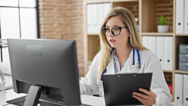 Mujer joven rubia médico utilizando computadora lectura informe médico en la clínica - Foto, imagen