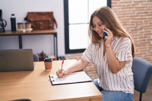 Młoda blondynka biznes pracownik mówi na smartfonie pisanie na schowku w biurze - Zdjęcie, obraz