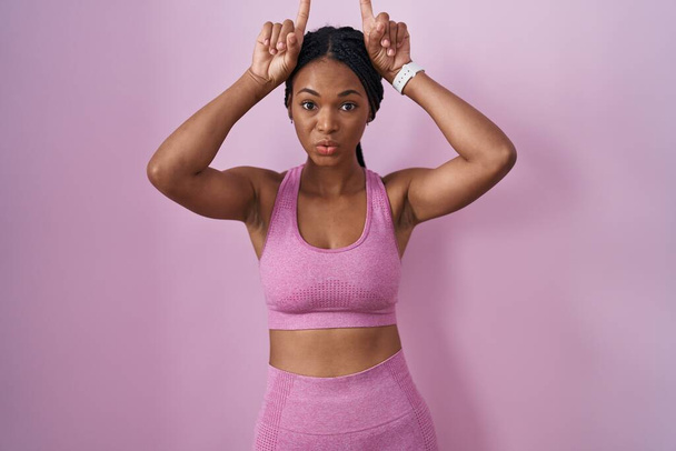 アフリカ系アメリカ人女性,ピンクの背景の上にスポーツウェアを着用し,頭の上に頭の上に指で面白いジェスチャーをする  - 写真・画像