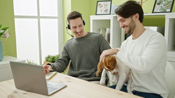 Dois homens casal usando laptop e fones de ouvido sentado na mesa com o cão na sala de jantar - Foto, Imagem