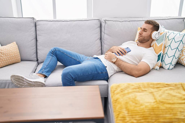 Młody biały mężczyzna słuchający muzyki leżącej na kanapie w domu - Zdjęcie, obraz