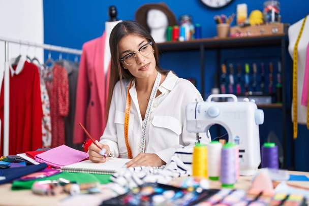 Young beautiful hispanic woman tailor drawing on notebook at clothing factory - Photo, Image