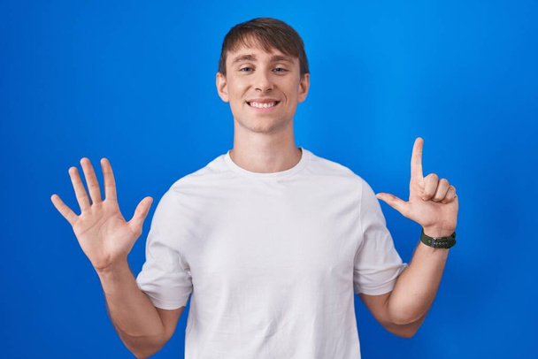 Běloch blond muž stojící nad modrým pozadím ukazuje a ukazuje s prsty číslo sedm a zároveň se usmívá sebevědomě a šťastně.  - Fotografie, Obrázek