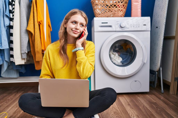 fiatal szőke nő használ laptop beszél okostelefon vár mosógép a mosókonyhában - Fotó, kép