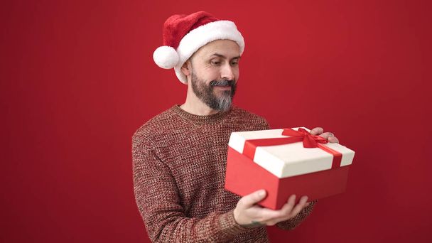 młody łysy człowiek sobie Boże Narodzenie kapelusz rozpakowywanie prezent na izolowane czerwone tło - Zdjęcie, obraz