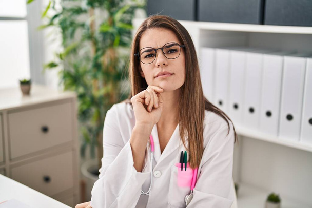 Young beautiful hispanic woman doctor speaking with serious expression at clinic - Photo, image