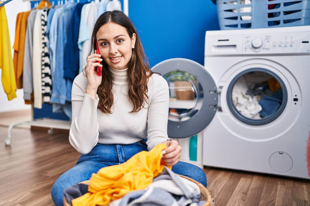 Joven hermosa mujer hispana sonriendo confiada hablando en el teléfono inteligente en la sala de lavandería - Foto, Imagen