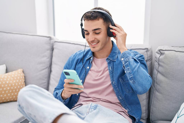Fiatal spanyol férfi zenét hallgat otthon, a kanapén ülve. - Fotó, kép