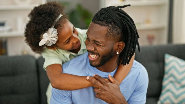 Африканский американец отец и дочь улыбаются уверенно обнимая друг друга сидя дома на диване - Фото, изображение