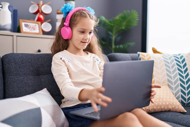 Menina caucasiana adorável usando laptop e fones de ouvido sentado no sofá em casa - Foto, Imagem